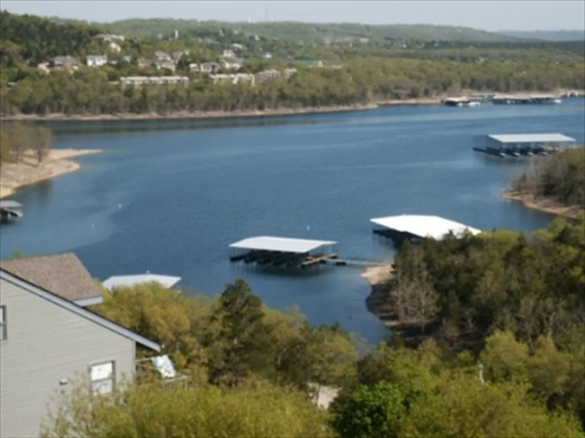 A tranquil lakeside scene showing calm water, boathouses, surrounding trees, and distant hills. The setting sun casts warm hues across the landscape, and the environment hints at recreational activities such as boating and fishing.