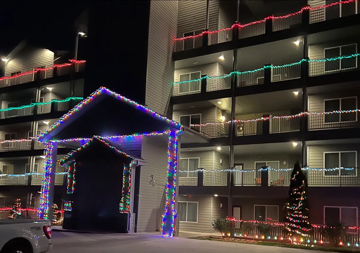 The image showcases the building's exterior decorated with festive lights, an inviting entrance awning, and warmly lit balconies. There's a Christmas tree visible near the entrance, suggesting a communal, celebratory atmosphere.