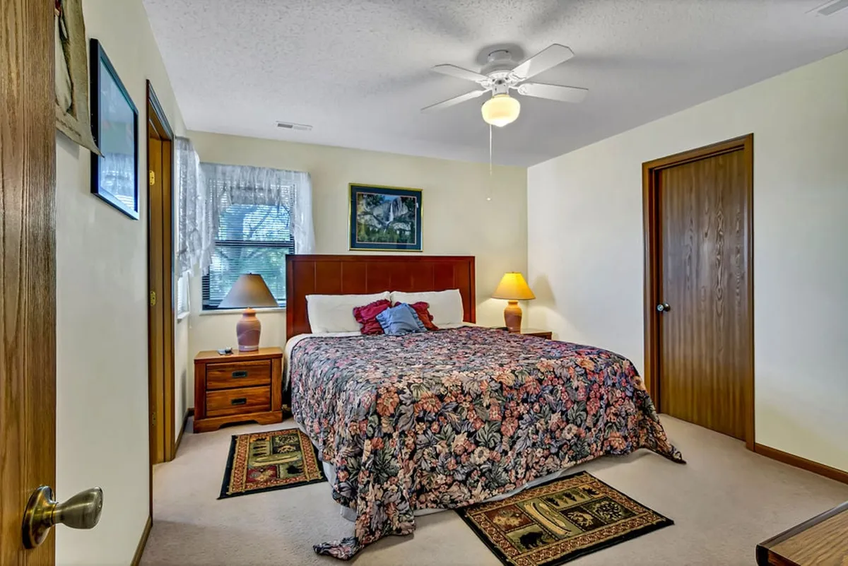 A cozy bedroom featuring a bed with floral bedding, a wooden nightstand topped with a lamp, and framed art on the wall. The natural light streaming in creates a peaceful atmosphere ideal for restful sleep and morning relaxation.