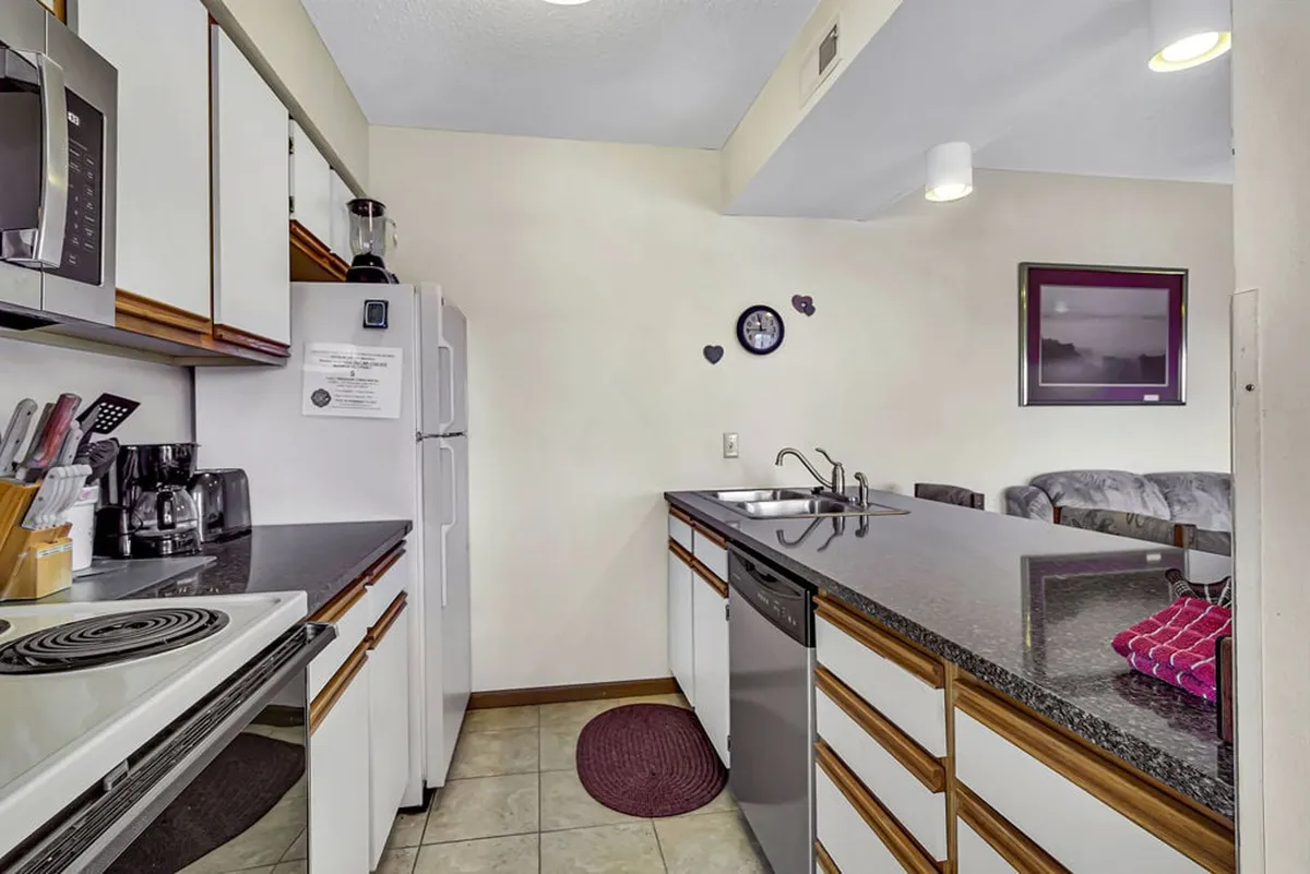This image highlights the kitchen area with a prominent granite counter, stainless steel appliances, colorful dish towels adding a retro flair, and a glimpse of a cozy couch in the background. The open layout invites group cooking, entertaining, and socializing.
