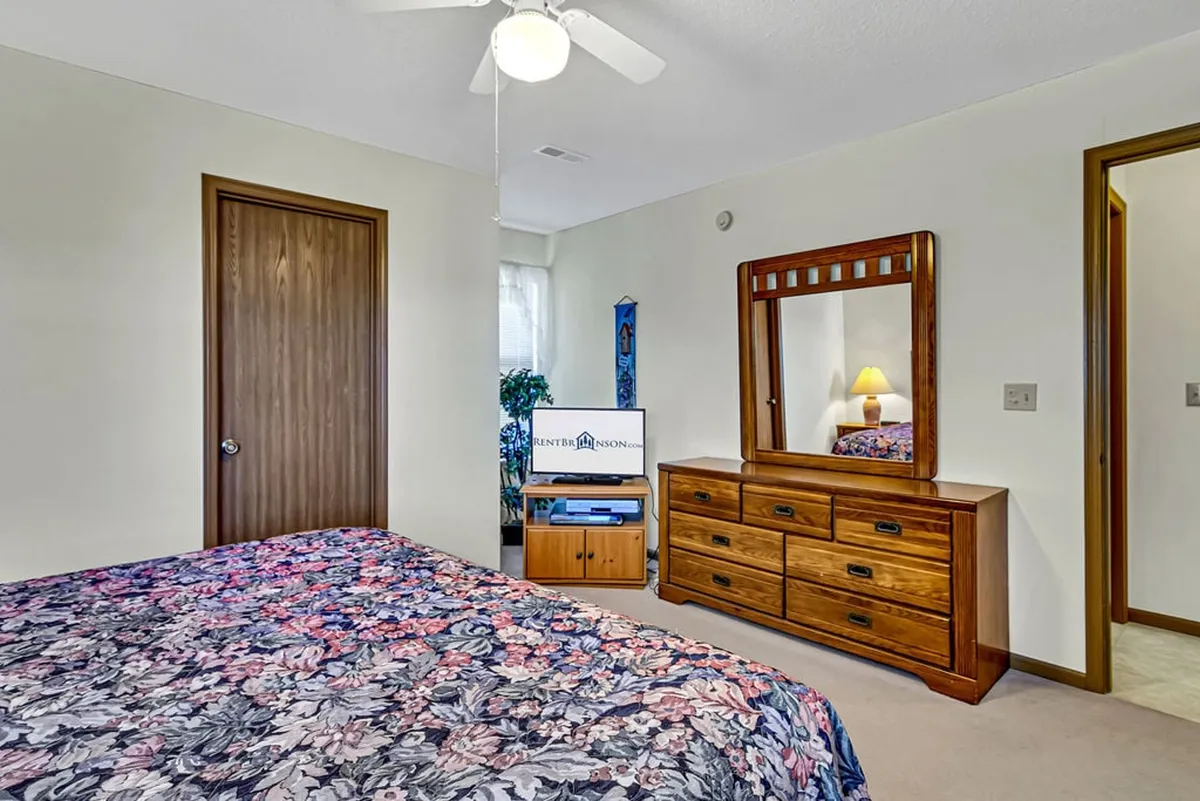 A cozy bedroom with a floral patterned bedspread, wooden dresser, mirrored vanity, and an indoor plant, featuring soft natural light filtering in. The room blends vintage touches with modern comfort, inviting relaxation and quiet moments.