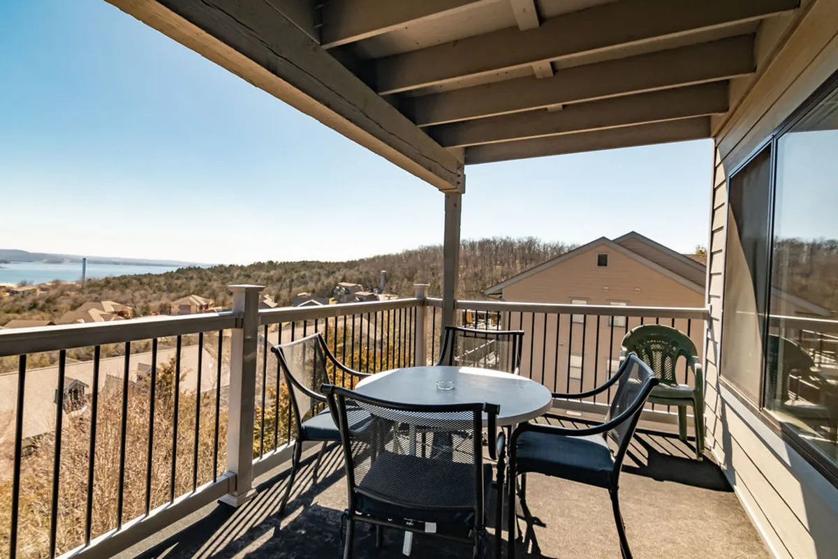 A sheltered outdoor deck featuring a dining table and chairs, set against a backdrop of lush landscape and railings that frame a panoramic lake view. Soft morning or evening light highlights the inviting space, suggesting it’s well-suited for coffee, meals, or relaxation while enjoying the views.