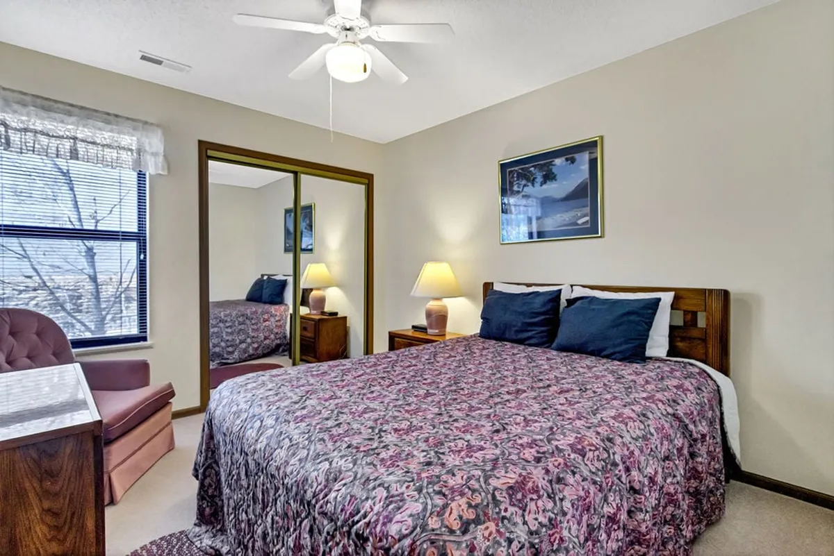 A welcoming bedroom with a patterned comforter on the bed, mirrored closet doors, a nightstand with a reading lamp, and framed artwork adorning the wall. The ambiance is both cozy and bright, suggesting a space for relaxation and comfort.