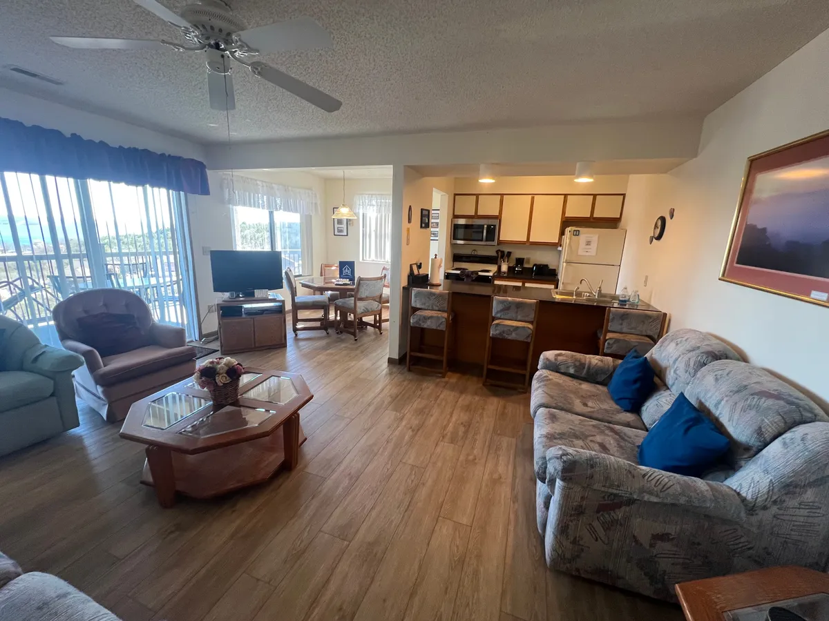 The image showcases an inviting living room with a plush sofa adorned with blue cushions, a glass coffee table perfect for game nights, and a clear view of the adjacent dining area with chairs. A TV setup is visible, emphasizing comfort and entertainment, while large windows fill the space with natural light.
