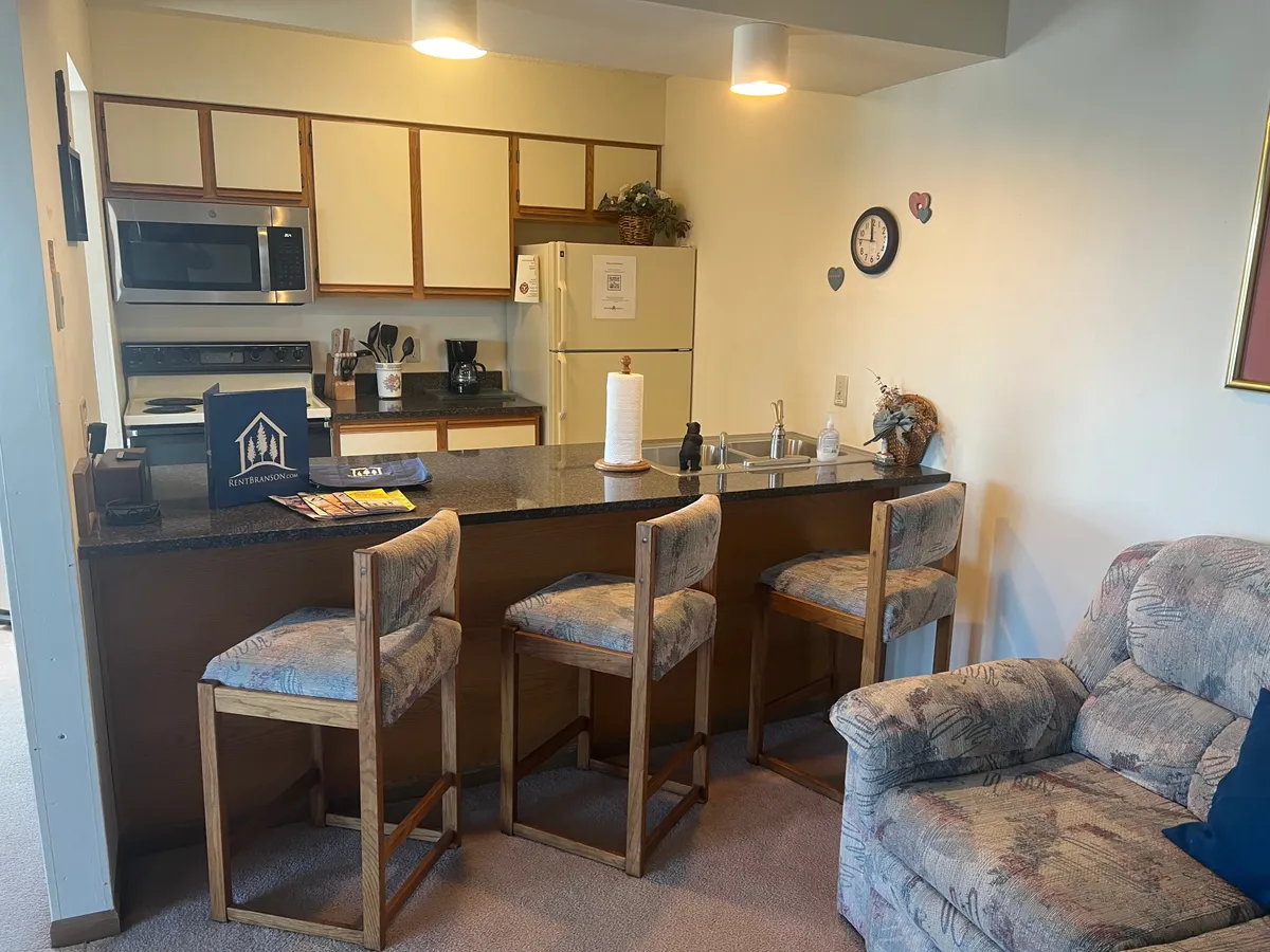 A cozy kitchen and living area featuring a granite countertop with bar stools, a comfortable couch, and a decorative wall clock. The space exudes a warm, vintage-inspired charm and is well-suited for both entertaining and quiet mornings.